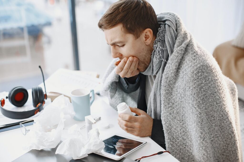 A white man in his twenties or thirties wrapped in a blanket. There are tissues on his desk and a mug, he's taking some tablets.