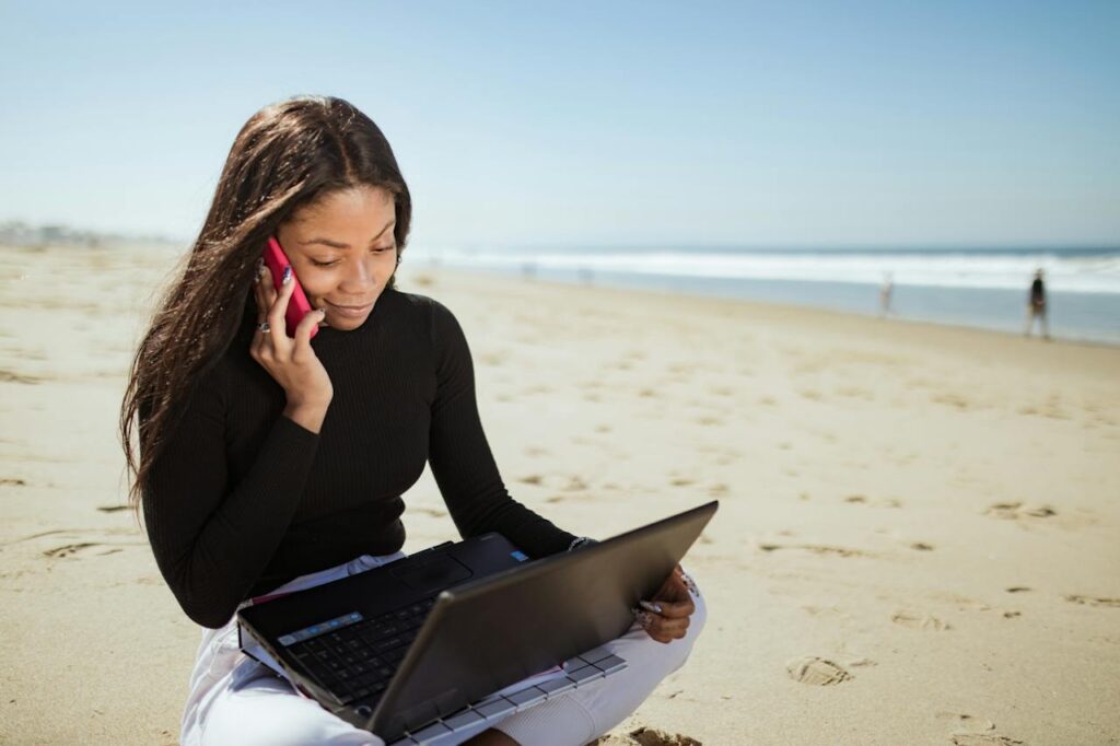 A black woman with long brown hair wearing white trousers and a black long-sleeved top sat on the beach with a pink cell phone in her hand and a laptop on her lap.
