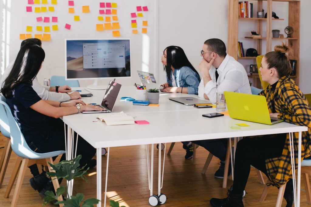 five colleagues are sat at a desk looking at a screen, some of them have their own laptops open. Behind the screen is a board with post-it notes