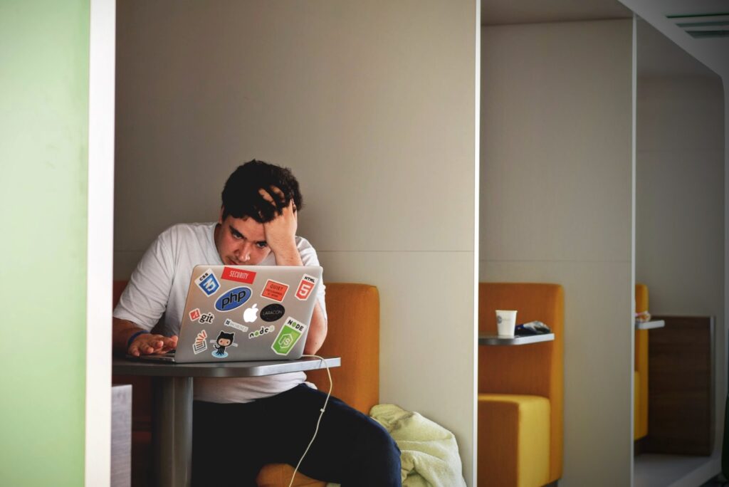 A man sat in a cubicle with a laptop covered in stickers. He has his head in his hand and looks frustrated