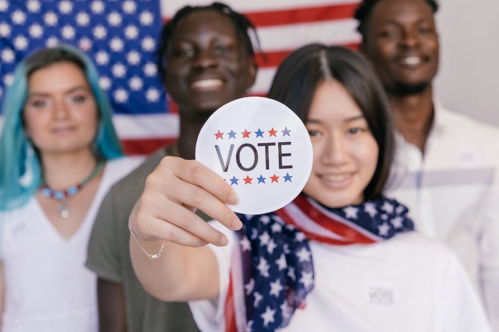 4 multiracial young people stood in front for the American flag. 2 men, 2 women. In the front is a south asian woman holding a vote sticker.