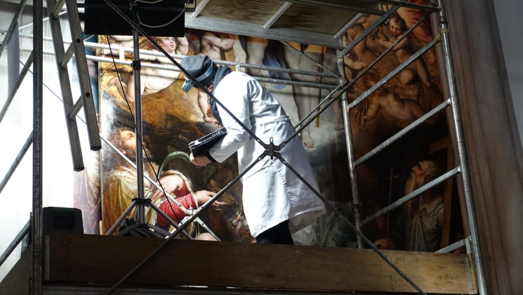A restorer standing on scaffold in front of a painting.