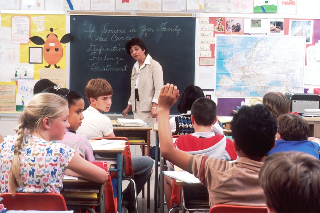 A teacher standing in front of a blackboard, in a classroom full of children.