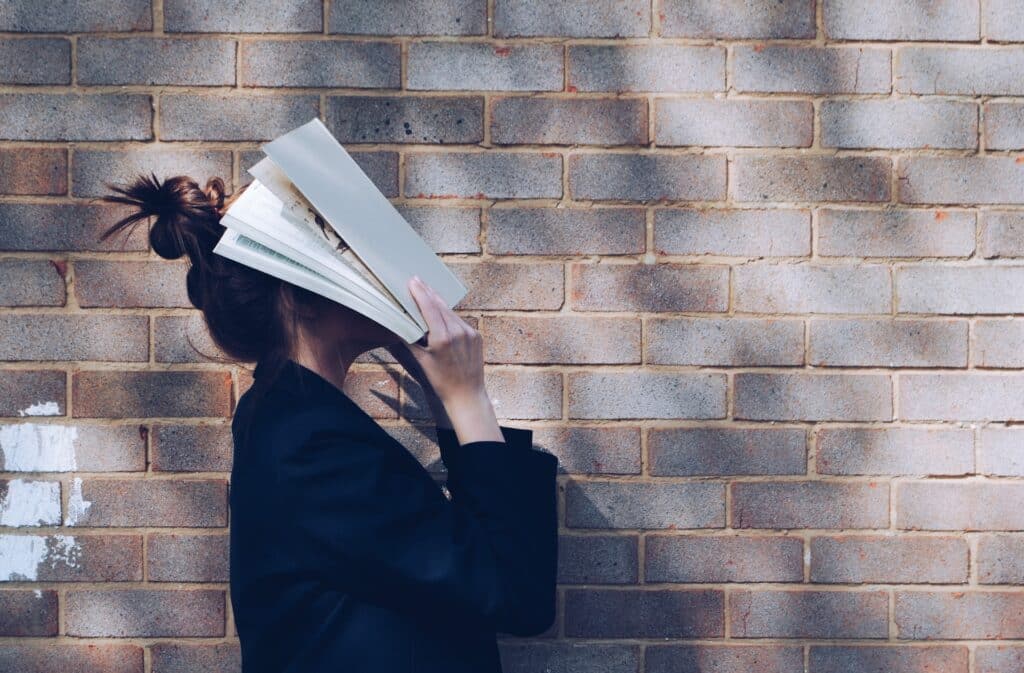 A woman standing in front of a brick wall, holding a book up to cover her face.